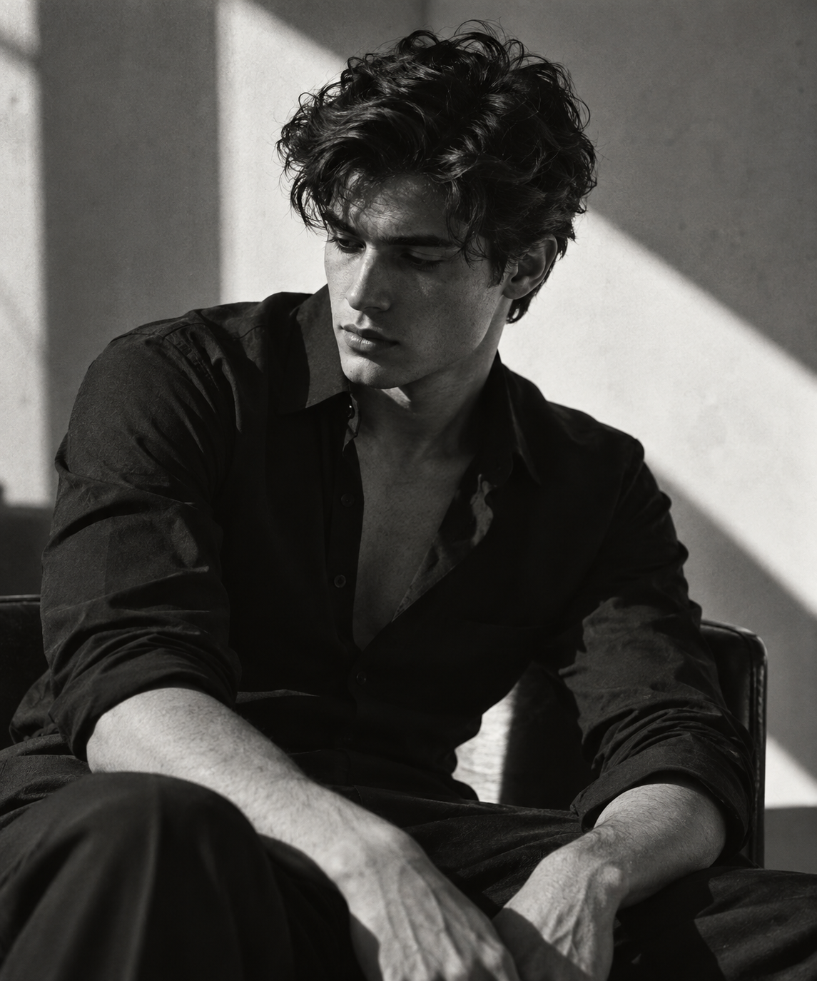 Black-and-white editorial portrait of a seated young man in an open black shirt, with a diagonal beam crossing the studio wall.